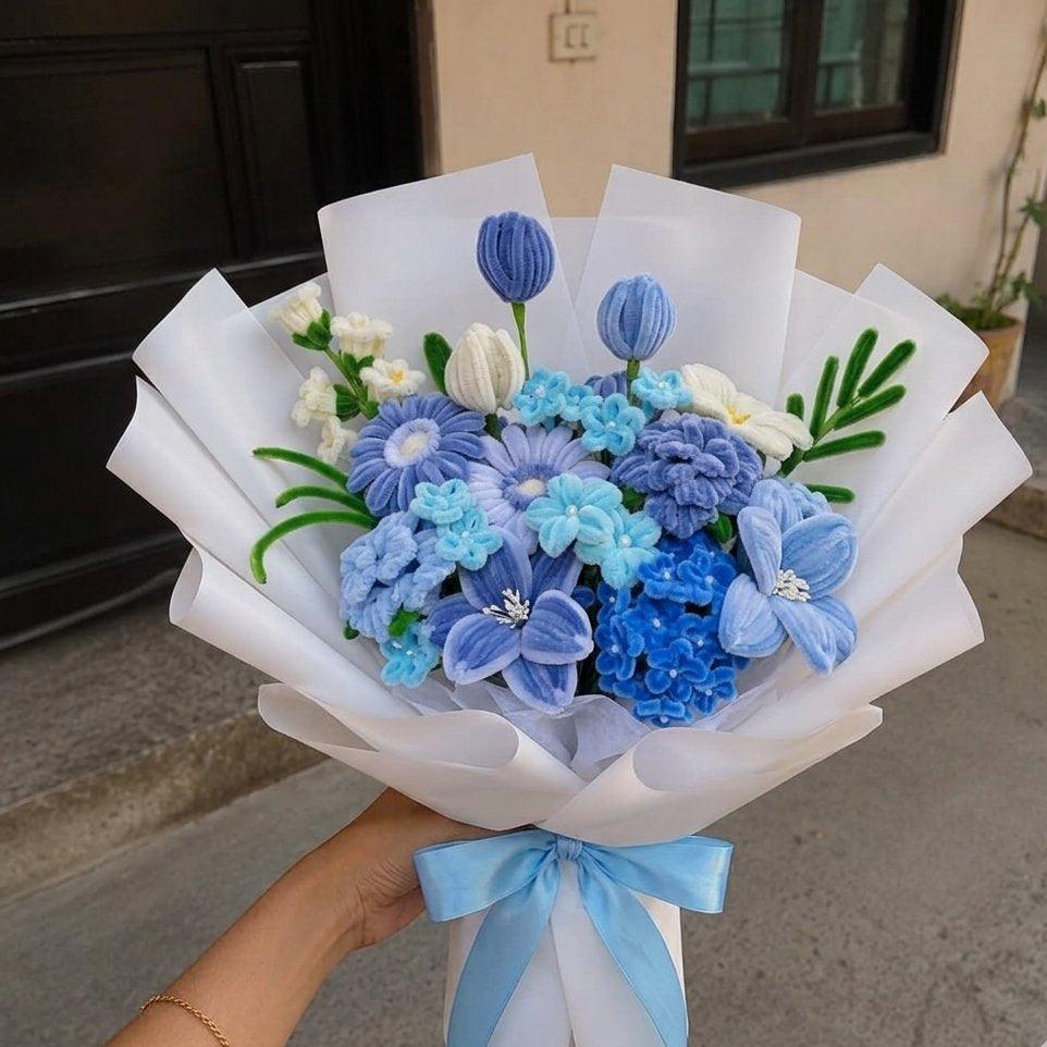 A hand holding a handcrafted bouquet of blue and light blue artificial flowers wrapped in white paper with a light blue ribbon, set against an outdoor background.