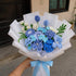 A hand holding a handcrafted bouquet of blue and light blue artificial flowers wrapped in white paper with a light blue ribbon, set against an outdoor background.